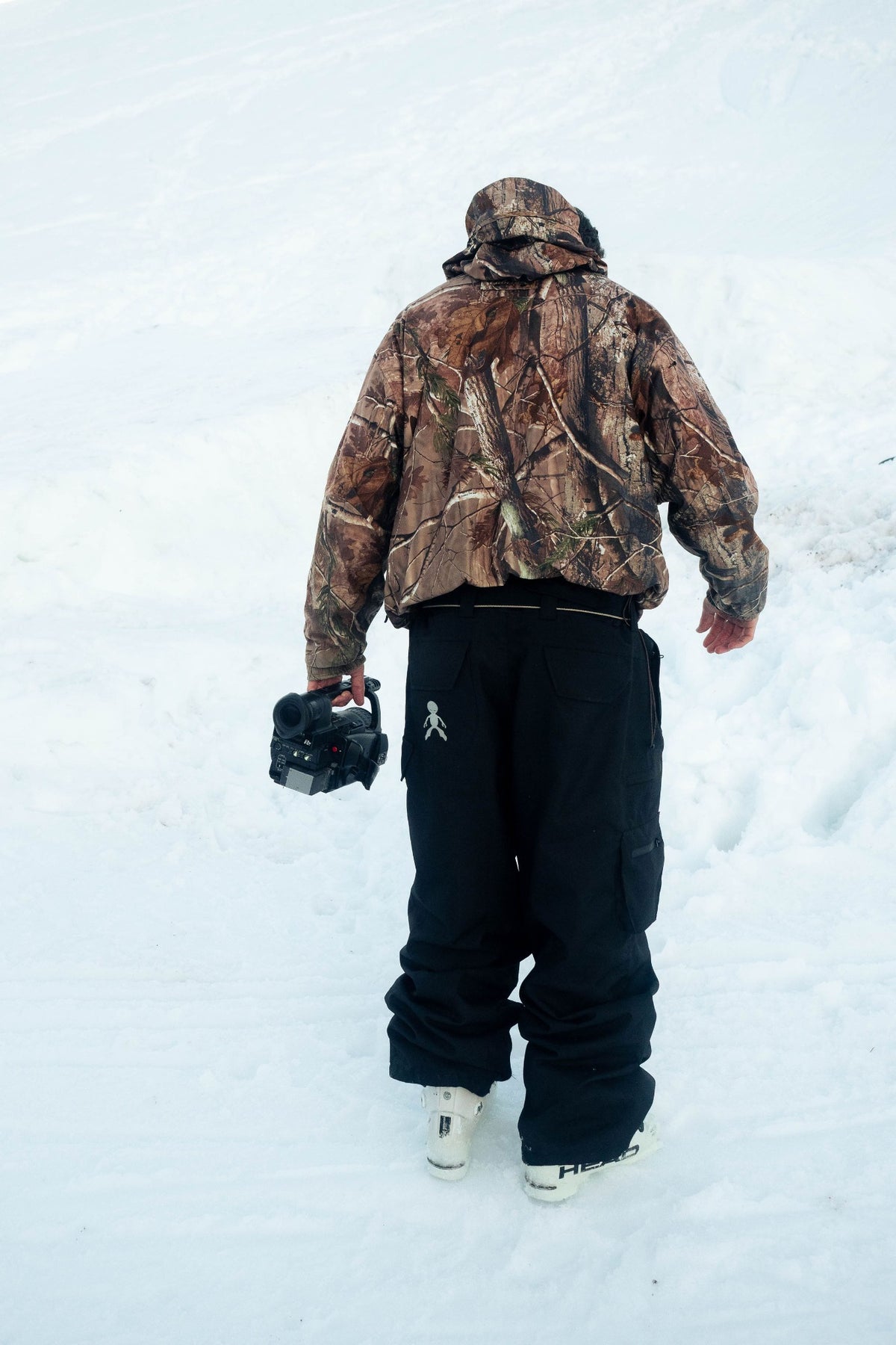 Skier wearing black Steeve ski pants and camouflage jacket walking through snow with a camera